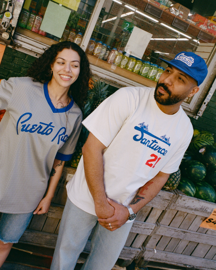 Two people stand by a fruit stand. The woman wears a gray Puerto Rico shirt; the man wears an Ebbets Field Puerto Rico 1958 National Team Mesh V-Neck Jersey styled with “Santurce 21” and a blue cap. Watermelons and drinks are in the background.