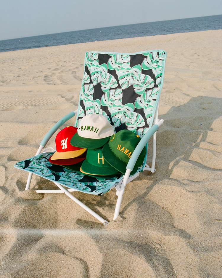 A beach chair with green leaf patterns sits on the sand, displaying four baseball caps—including the EFF Hawaii Islanders 1986 Authentic Wool Ballcap—with the ocean and sky as a serene backdrop.