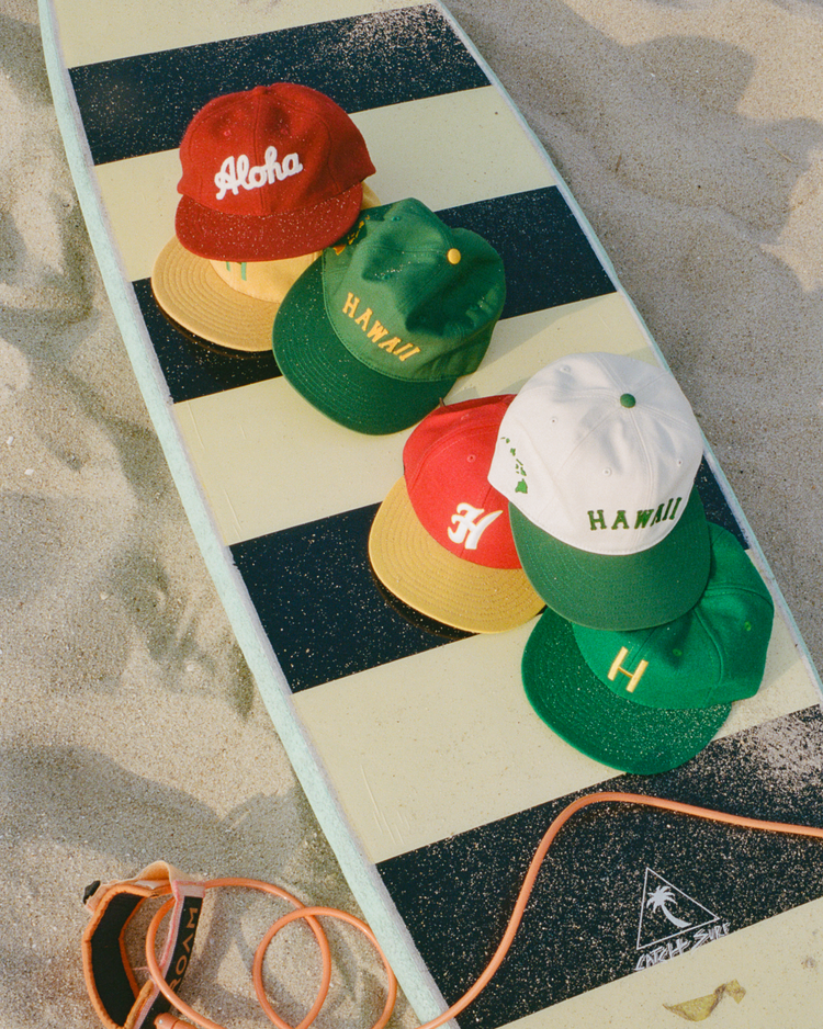 Five Ebbets Field cotton twill hats, including the Hawaii Islanders (PCL) Ballcap, are displayed on a striped surfboard at the beach. The hats show words like Aloha and Hawaii, with an orange surf leash beside the board.