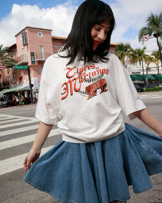 A young woman stands on a city street, eyes closed and smiling. She wears the Ebbets Field Marianao Tigres Short Sleeve Graphic Tee - Cuban League with a blue denim skirt. Palm trees and pastel buildings are in the background.