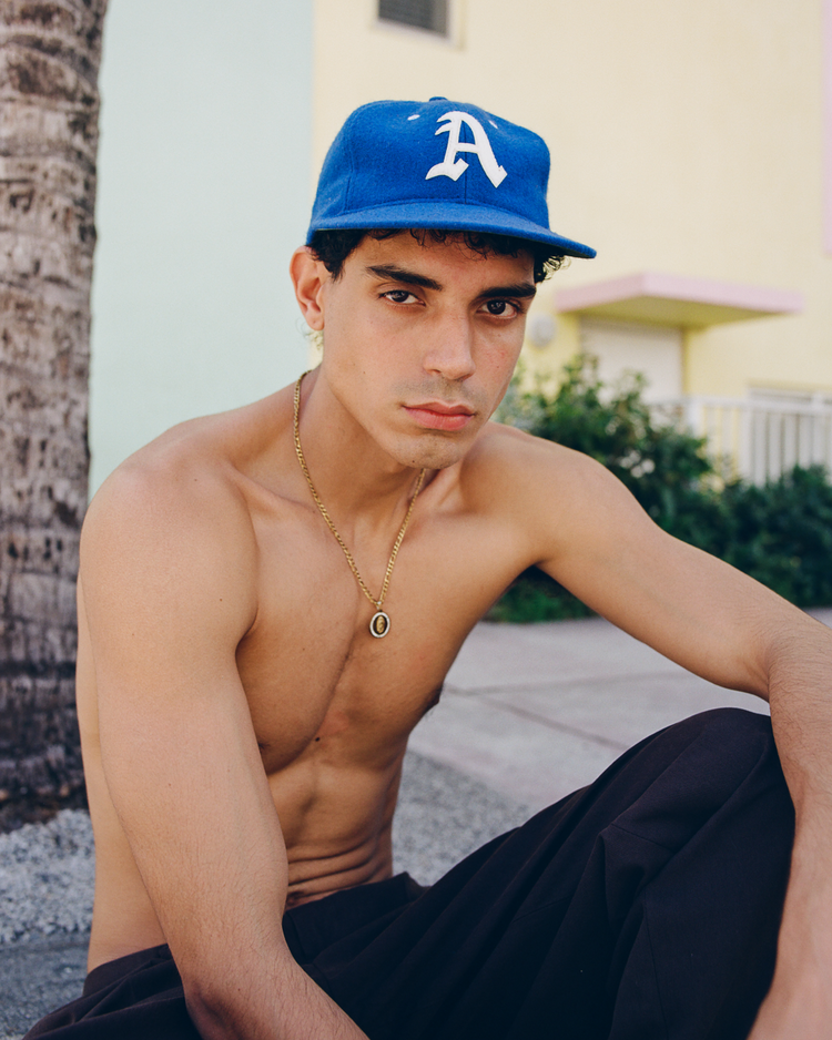 A young man sits outdoors, shirtless, wearing an Ebbets Field Almendares Alacranes 1947 Authentic Wool Ballcap and a gold necklace. He looks seriously at the camera, with greenery and a pastel building behind him.