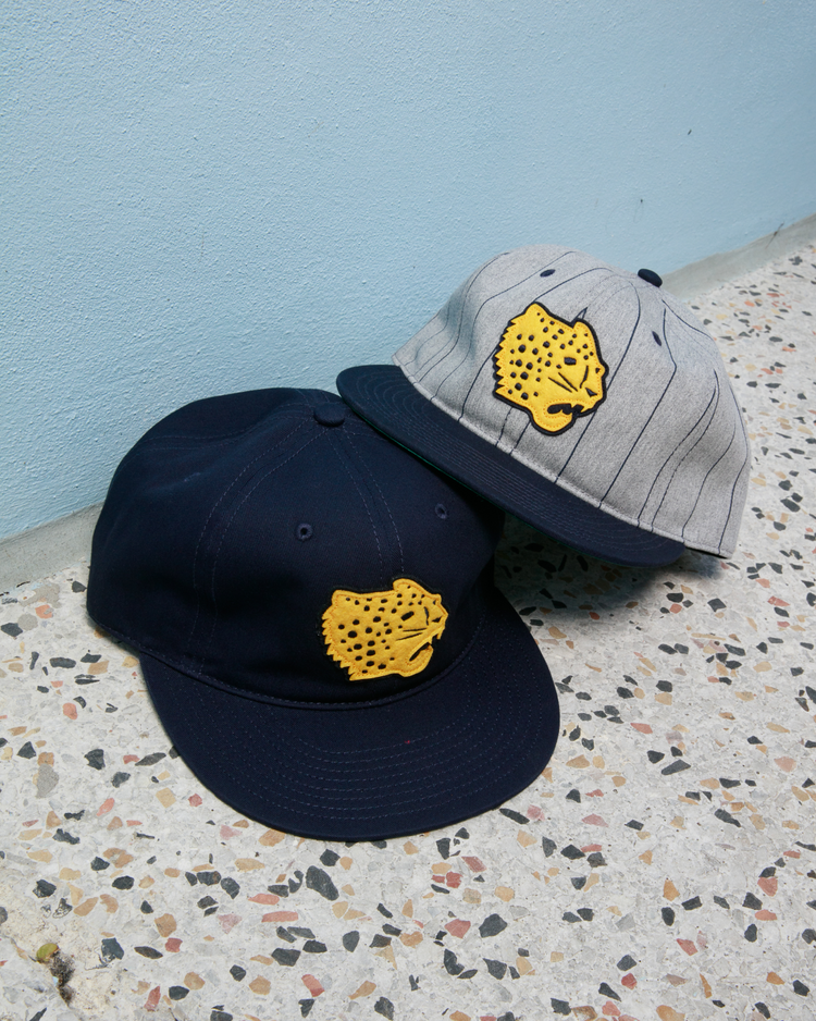 Two Ebbets Field Santa Clara Leopardos 1925 Cotton Twill Ballcaps sit on a speckled floor by a light blue wall—one navy, one striped light gray—both featuring the classic yellow cheetah head logo from Cuban baseball lore.