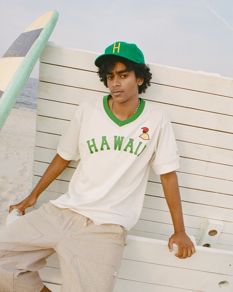 A young man in an EFF Hawaii Islanders 1961 Authentic Wool Ballcap and a white HAWAII jersey sits on a white wooden beach bench, surfboard and sand behind him.