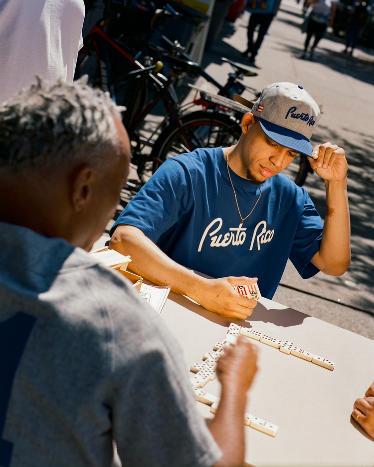 Two men play dominoes at an outdoor table on a sunny day. One wears a Roberto Clemente shirt and an Ebbets Field Puerto Rico 1958 National Team Cotton Twill Ballcap, holding domino pieces. Bicycles and pedestrians pass by in the background.