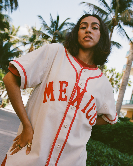 A woman with shoulder-length dark hair poses outdoors in an Ebbets Field Diablos Rojos del Mexico Cotton Twill Baseball Jersey, cream-colored with red trim and 