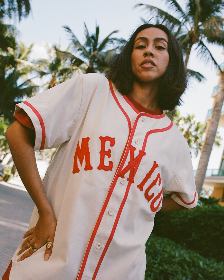 A woman with shoulder-length dark hair poses outdoors in an Ebbets Field Diablos Rojos del Mexico Cotton Twill Baseball Jersey, cream-colored with red trim and 