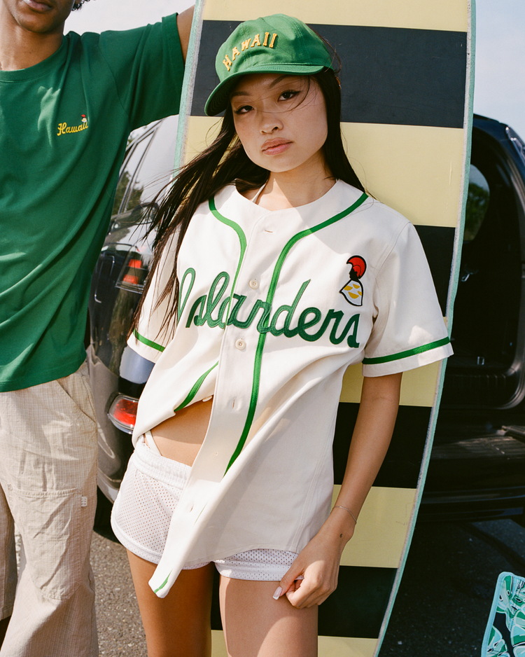 A woman wears an Ebbets Field Hawaii Islanders (PCL) Cotton Twill Baseball Jersey, white shorts, and a green cap. She stands by a striped surfboard next to someone in a green “Hawaii” shirt with a car in the sunny background.