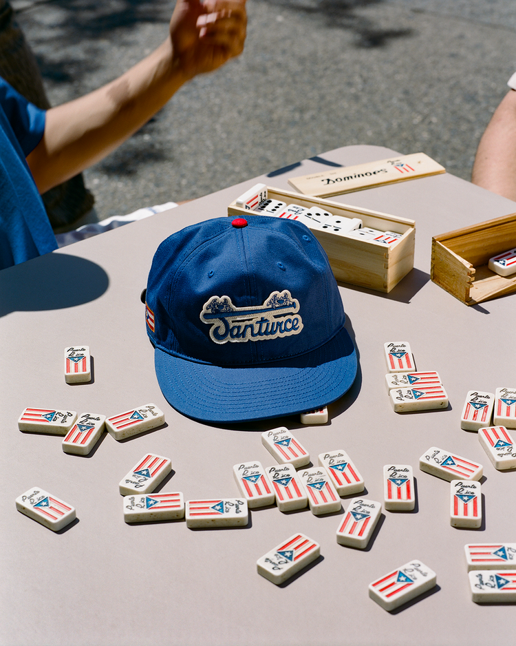 An Ebbets Field Santurce Cangrejeros Cotton Twill Ballcap with Santurce embroidered rests on a table with Puerto Rican flag dominoes, as two people play dominoes outdoors, evoking the lively spirit of Puerto Rican baseball.
