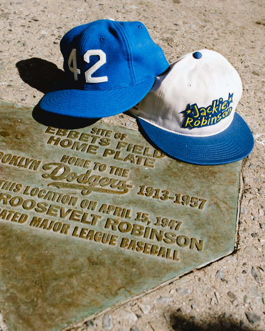 Two Ebbets Field Jackie Robinson Comic Book Hero Cotton Twill Ballcaps rest on a plaque marking Ebbets Field’s home plate, honoring Jackie Robinson and the Brooklyn Dodgers. One ballcap reads “42,” the other says “Jackie Robinson.”.