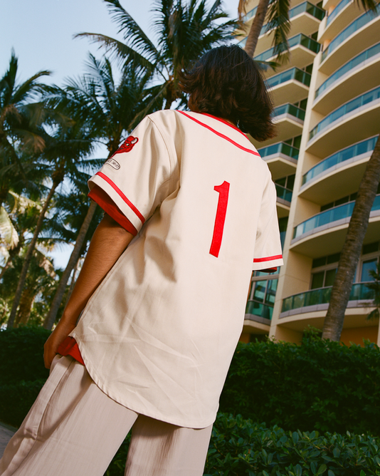 A person stands outdoors near palm trees and a modern building, wearing an Ebbets Field Diablos Rojos del Mexico Cotton Twill Baseball Jersey in cream with red trim and a large red number 1 on the back. The person's face is not visible.
