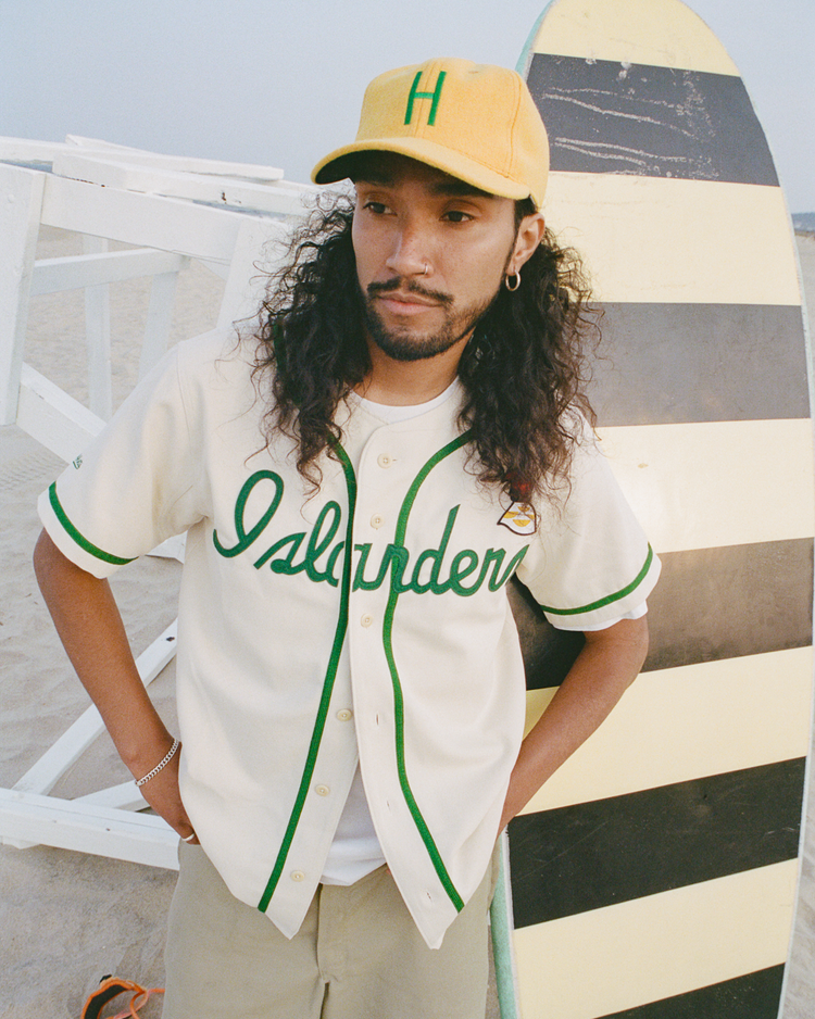 A bearded person with long curly hair wears a yellow cap and an Ebbets Field Hawaii Islanders (PCL) Cotton Twill Baseball Jersey, standing by a striped surfboard on the sandy beach.