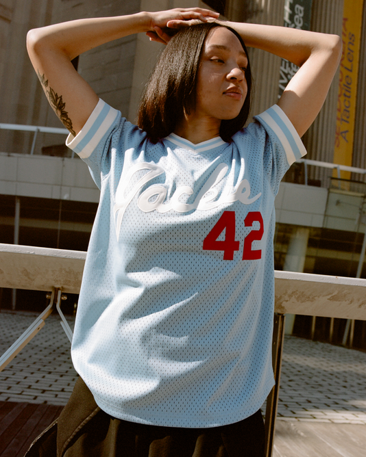 A woman with straight dark hair stands outdoors in front of a modern building, arms raised behind her head, wearing the Ebbets Field Jackie Robinson Powder Blue V-Neck Mesh Jersey with white and red lettering and the number 42.