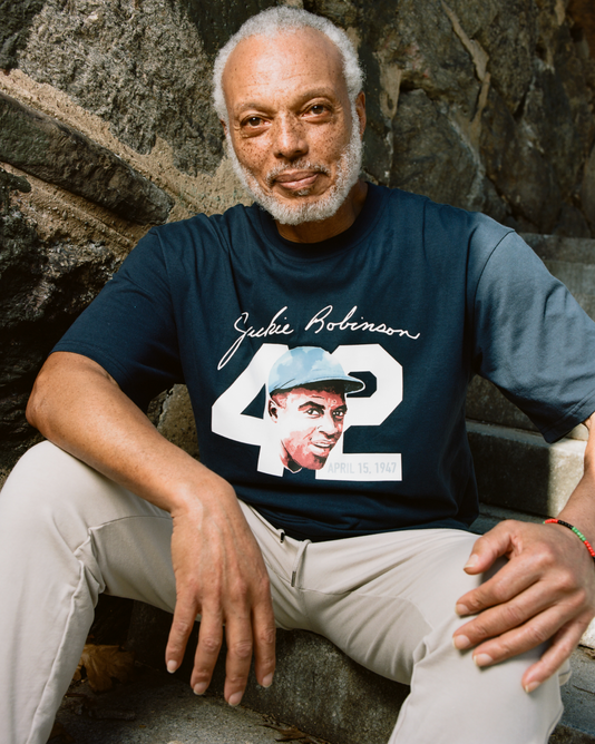 An older man with gray hair and a beard sits on outdoor steps by a stone wall, wearing the Ebbets Field Jackie Robinson Watercolor Portrait Short Sleeve Graphic Tee. He looks at the camera with a relaxed expression, one arm resting on his knee.