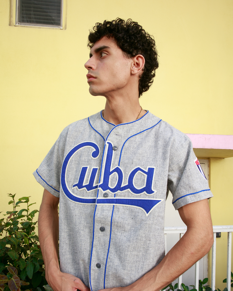 A young man with curly dark hair wears the EFF Cuba National Team 1947 Authentic Wool Road Baseball Jersey, standing before a yellow wall and looking to the side, with plants and part of a white railing behind him.
