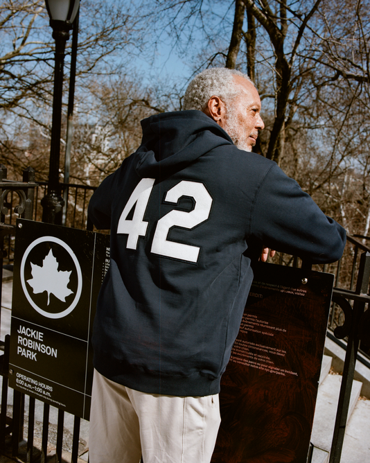 A gray-haired man wears an Ebbets Field Jackie Robinson All-Stars Heavyweight Cotton Terry Pullover Hoodie, featuring number 42 on the back, as he stands by signs at Jackie Robinson Park with leafless trees in view.