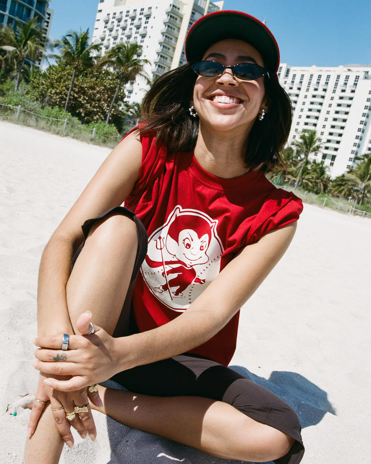 A woman in a red cap, sunglasses, and an Ebbets Field Diablos Rojos del Mexico Short Sleeve Graphic Tee with a cartoon devil sits smiling on a sandy beach, with palm trees and tall buildings behind her.
