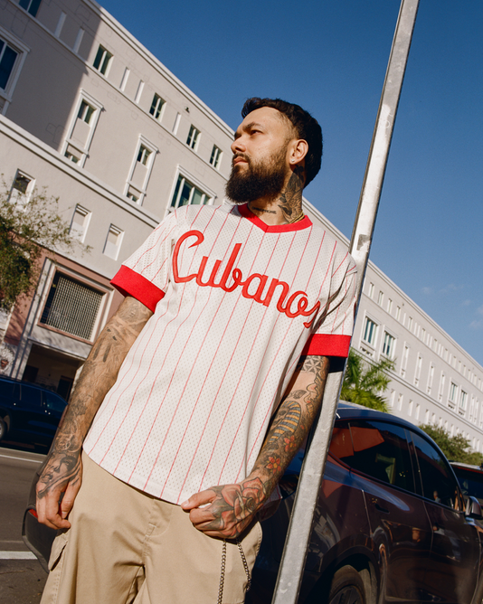 A bearded, tattooed man wearing the Ebbets Field Havana Cubans Mesh V-Neck Jersey and beige shorts leans against a pole on a city street, with buildings and parked cars in the background.