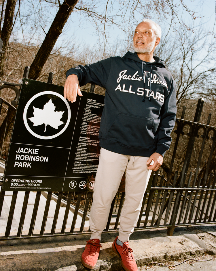 An older man stands by a Jackie Robinson Park sign, wearing light pants, red shoes, and the Ebbets Field Jackie Robinson All-Stars Heavyweight Cotton Terry Pullover Hoodie, with trees and a black metal fence behind him.