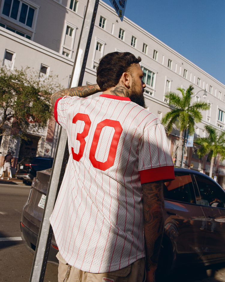 A tattooed man wears the Ebbets Field Havana Cubans Mesh V-Neck Jersey, featuring red and white pinstripes and the number 30, standing on a sunny street lined with parked cars and palm trees.