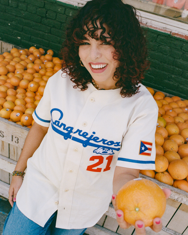 A smiling person with curly hair wears the Ebbets Field Santurce Cangrejeros Cotton Twill Baseball Jersey, featuring the Puerto Rican flag and number 21, standing by oranges and holding one toward the camera.