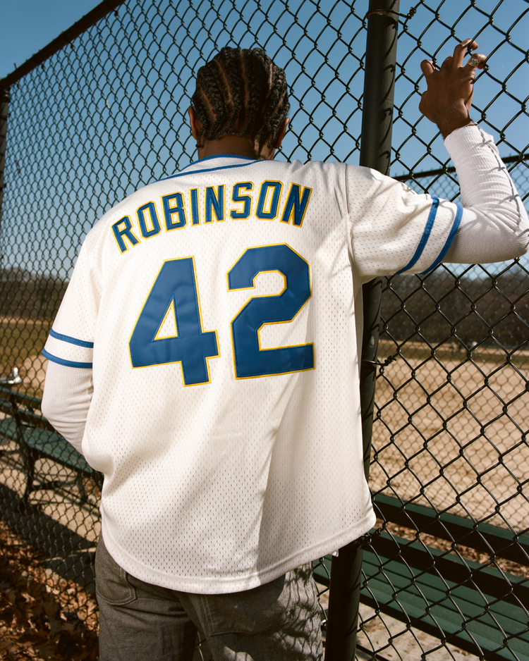A person with braided hair stands by a chain-link fence, wearing an Ebbets Field Jackie Robinson Comic Book Hero Mesh Baseball Jersey. Blue stripes and the number 42 are visible on the back, with a baseball field in the background.