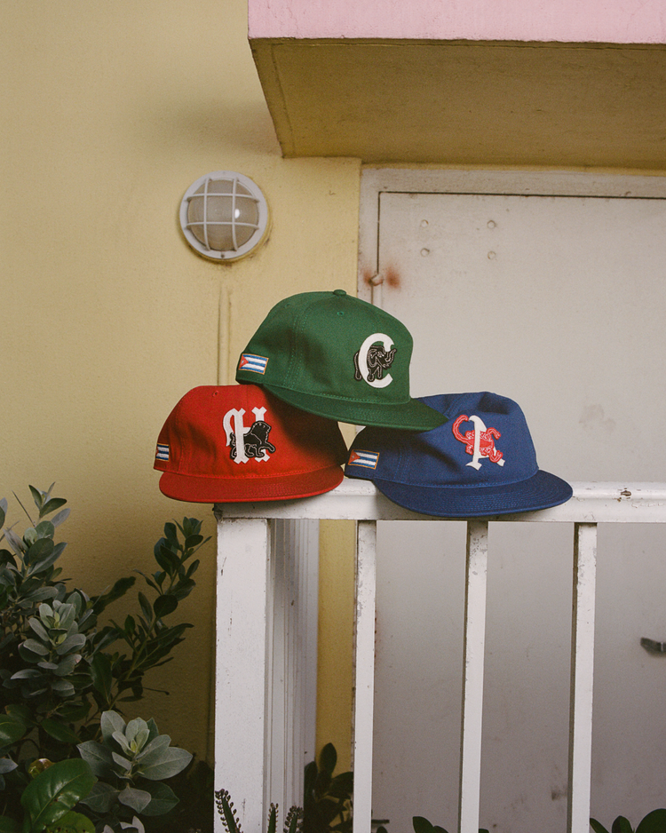 Three Cienfuegos Elefantes Cotton Twill Ballcaps by Ebbets Field, in red, green, and blue with unique Cuban baseball-inspired embroidery, sit on a white railing against a pastel yellow wall with plants and an outdoor light nearby.