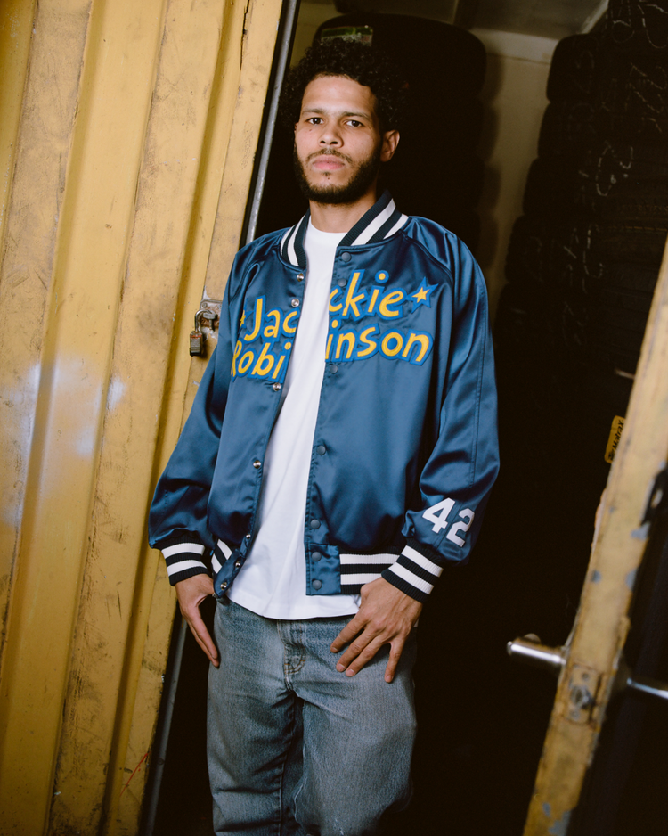 A man stands in a doorway wearing the Ebbets Field Jackie Robinson Comic Book Hero Cotton Lined Satin Varsity Jacket—blue with the number 42—over a white t-shirt and jeans, with yellow metal walls and stacked black tires in the background.