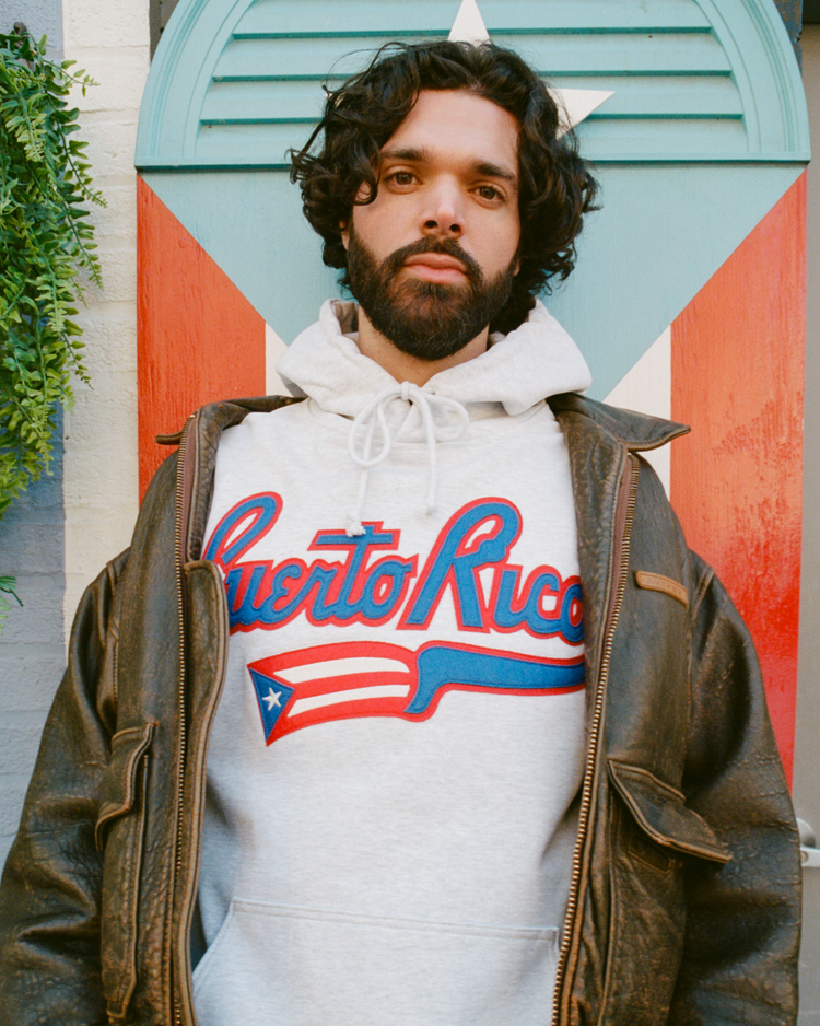 A man with curly hair and a beard wears an Ebbets Field Puerto Rico 1972 National Team Heavyweight Cotton Terry Pullover Hoodie and a brown leather jacket, standing before a wall painted with a Puerto Rican flag. Green plants hang to the left.