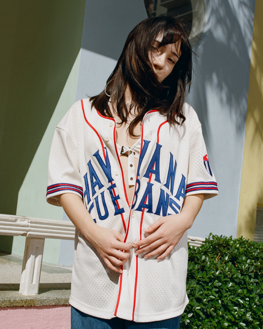 A woman stands outdoors in sunlight, wearing the Ebbets Field Havana Cubans Mesh Baseball Jersey. Her hair partly covers her face, with colorful buildings and greenery in the background.