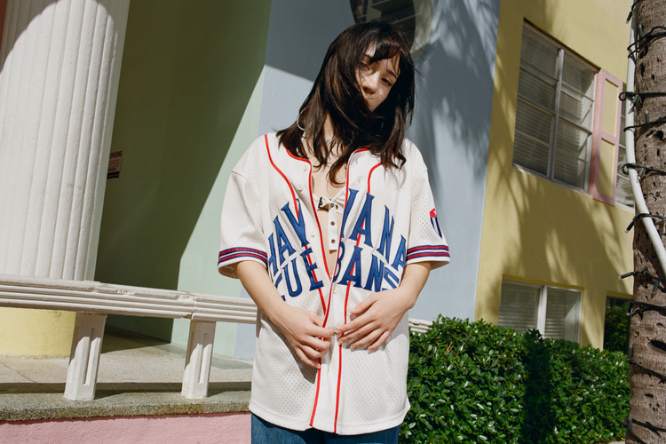 A woman stands outdoors in sunlight, wearing the Ebbets Field Havana Cubans Mesh Baseball Jersey. Her hair partly covers her face, with colorful buildings and greenery in the background.