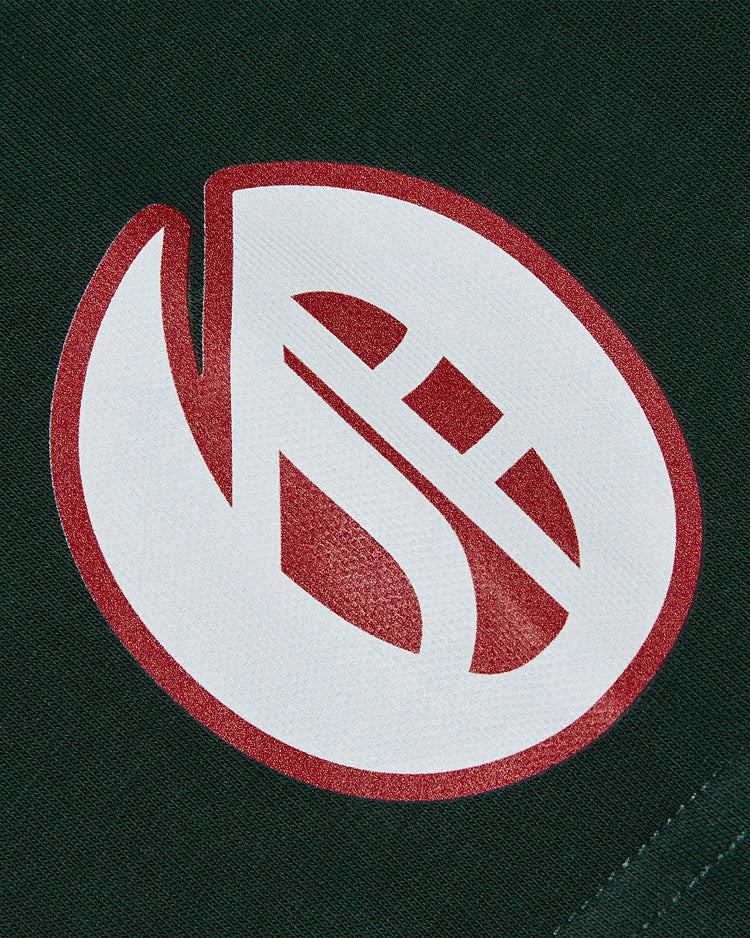 A close-up of the Oakland Oaks Ebbets Tee with Back Print by Ebbets Field shows a stylized white and red abstract logo—curved and diagonal lines in an oval shape—on dark green fabric.