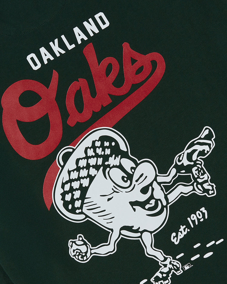 The Oakland Oaks Ebbets Tee by Ebbets Field is a dark green shirt with Oakland Oaks in white and red, a cartoon acorn with arms, legs, sneakers, and “Est. 1963” near the lower right. Features back print.