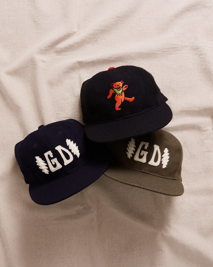 Two Ebbets Field wool broadcloth Grateful Dead Double Bolt ballcaps—one navy, one olive—rest next to a black Grateful Dead cap with an orange dancing bear, perfect for commemorating the 60th anniversary.