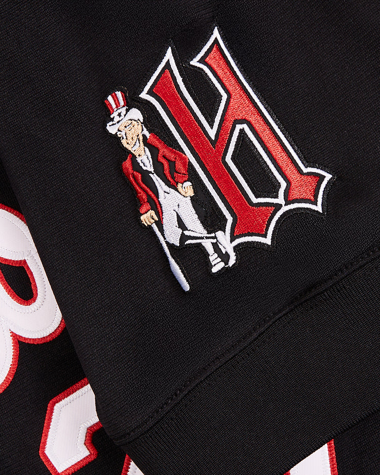 Close-up of black fabric from an EFF Vladimir Guerrero Harrisburg Senators V-Neck Jersey, showing a red and white letter H and an embroidered Uncle Sam in a top hat, red jacket, and cane. Part of another letter is also visible.