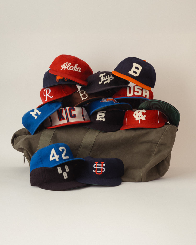 A green duffel bag holds colorful baseball caps, including the EFF Seattle Rainiers 1955 Authentic Wool Ballcap, each with unique letters, numbers, and logos in vibrant styles displayed against a plain background.