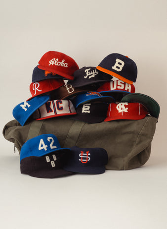 A pile of colorful baseball caps with various letters and logos stacked on top of a green duffel bag against a plain white background.