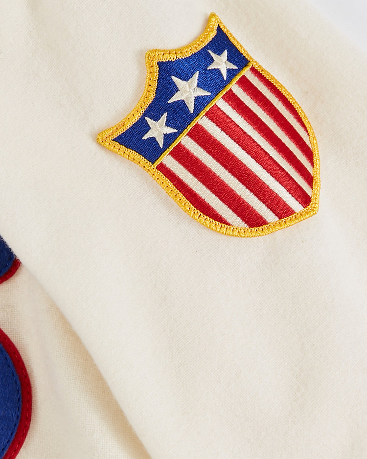 A close-up of the USA National Team 1936 Authentic Wool Baseball Jersey by Ebbets Field, showing cream-colored wool fabric and a shield patch with gold trim, blue top with three white stars, and red and white stripes below.
