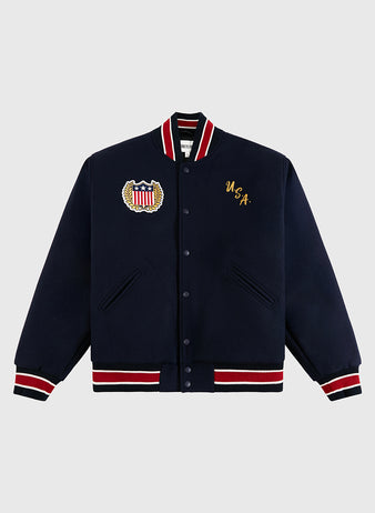 The Ebbets Field USA 1960 All-Wool Varsity Jacket in navy features red, white, and black striped trims, a vintage wool build, USA crest patch, and gold USA chest embroidery inspired by Olympic hockey gold moments.