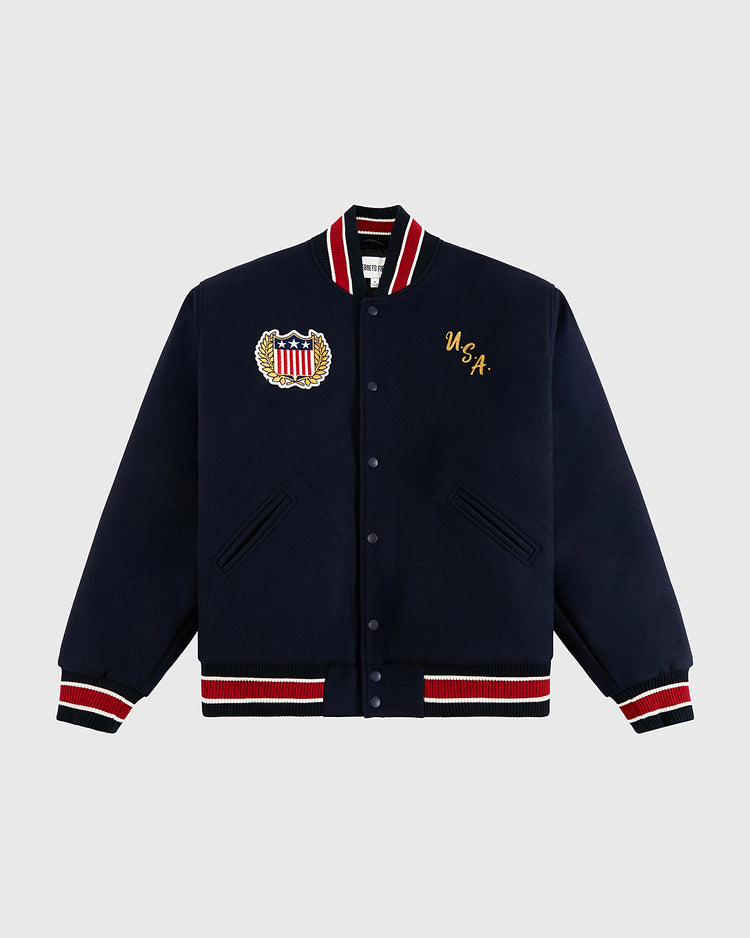 The Ebbets Field USA 1960 All-Wool Varsity Jacket in navy features red, white, and black striped trims, a vintage wool build, USA crest patch, and gold USA chest embroidery inspired by Olympic hockey gold moments.