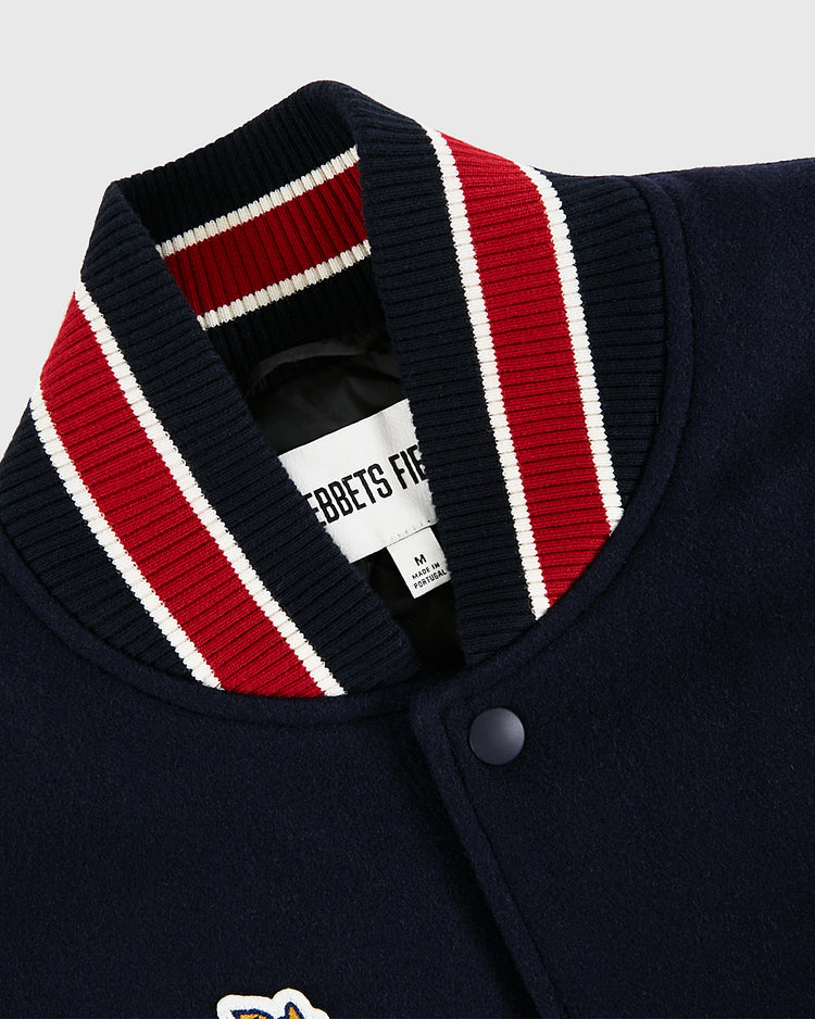 Close-up of a navy blue USA 1960 All-Wool Varsity Jacket collar by Ebbets Field, featuring red, white, and navy striped trim, snap button, and a visible inner label reading EBBETS FIELD in size M.