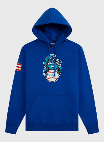 The Ebbets Field Santurce Cangrejeros Heavyweight Cotton Pullover Hoodie features a blue design with a cartoon crab holding a baseball on the front, red-and-white striped sleeve patch, front pocket, and drawstrings—Puerto Rican baseball-inspired.