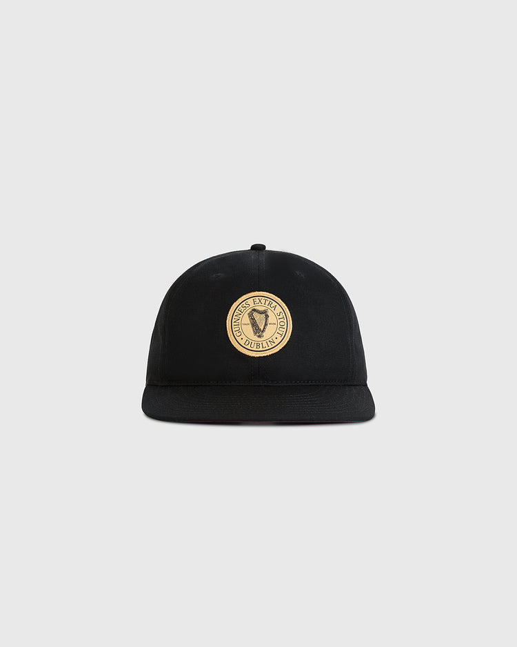 The Ebbets Field Guinness Extra Stout Patch Cotton Twill Ballcap is a black flat-brim cap with a round beige patch on the front, featuring 