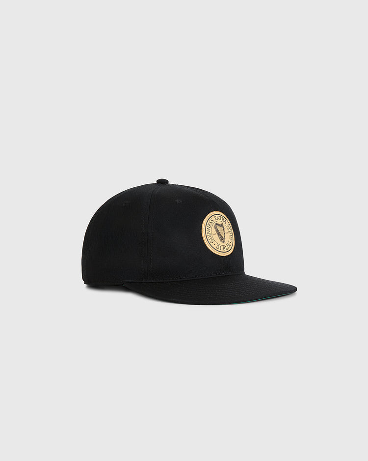 The Ebbets Field Guinness Extra Stout Patch Cotton Twill Ballcap is a black flat-brim hat with a round beige patch featuring stylized graphics and text, shown against a light gray background. Inspired by classic Ebbets Field design.