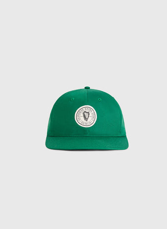 The Ebbets Field Guinness Extra Stout Patch Cotton Twill Ballcap is a green cap with a curved brim and a circular white patch featuring a black and white emblem on the front, styled after classic Ebbets Field designs. The cap is shown on a light gray background.
