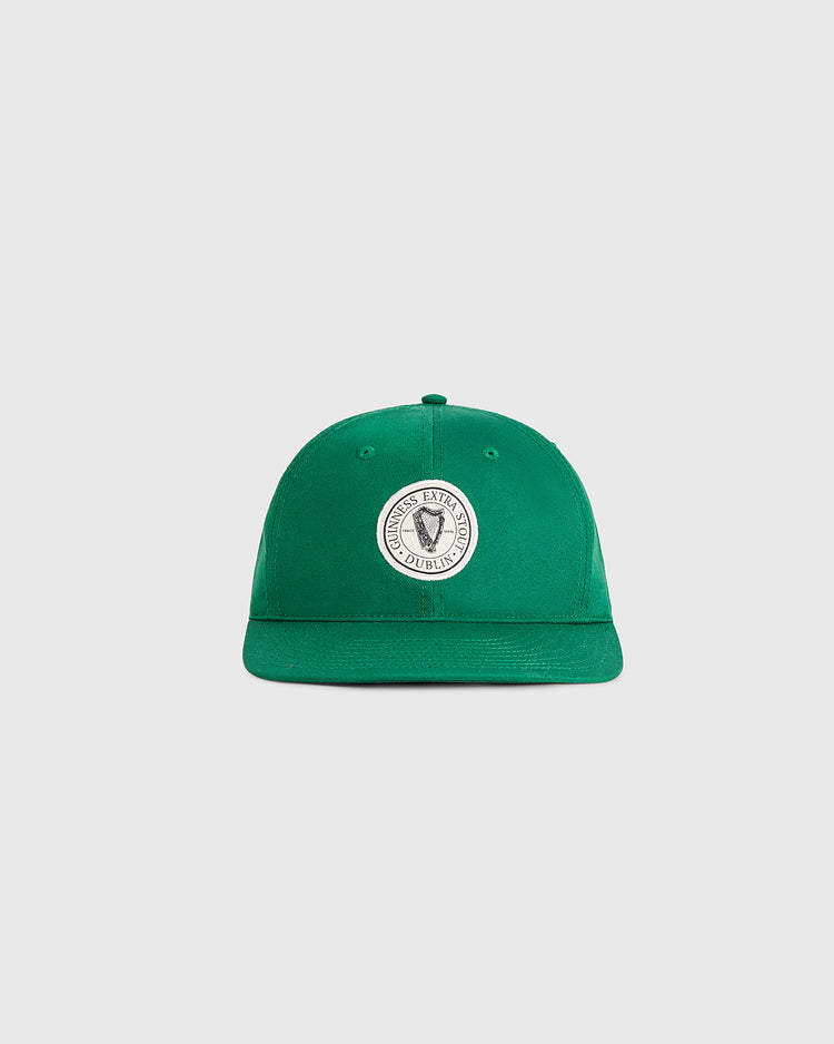 The Ebbets Field Guinness Extra Stout Patch Cotton Twill Ballcap is a green cap with a curved brim and a circular white patch featuring a black and white emblem on the front, styled after classic Ebbets Field designs. The cap is shown on a light gray background.