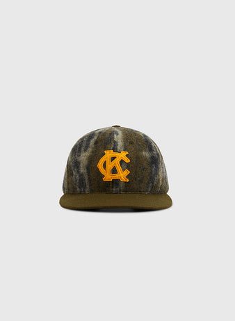 Front view of a KANSAS CITY MONARCHS 1945 MOSSY OAK cap by Ebbets Field, featuring a camouflage pattern, flat brim, and bold yellow embroidered AC logo on the front, set against a plain light gray background.