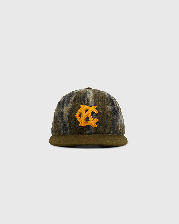 Front view of a KANSAS CITY MONARCHS 1945 MOSSY OAK cap by Ebbets Field, featuring a camouflage pattern, flat brim, and bold yellow embroidered AC logo on the front, set against a plain light gray background.