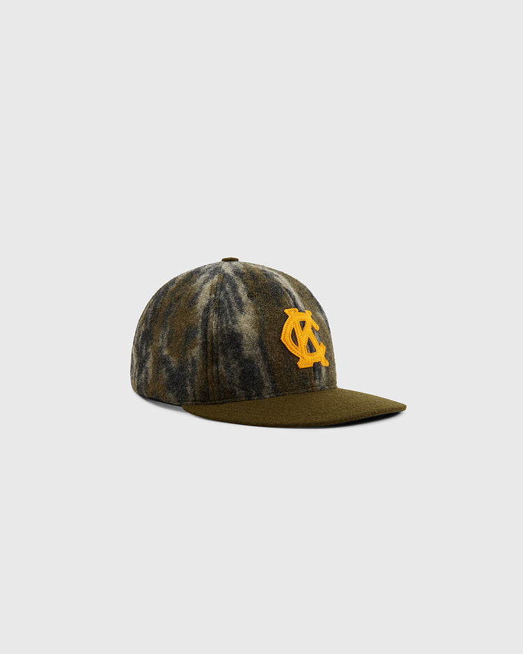 The Ebbets Field Mossy Oak x 1945 Kansas City Monarchs Ballcap features a green and brown camo pattern, flat brim, and a large yellow C and A logo, inspired by classic Negro Leagues hats, shown on a plain light gray background.