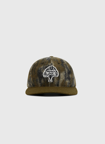 The Ebbets Field Mossy Oak x Ebbets Field Vintage Tree Logo Ballcap features a camouflage design, olive green brim, and black-and-white tree logo patch with “Mossy Oak” text. The cap is displayed centered on a plain light background.
