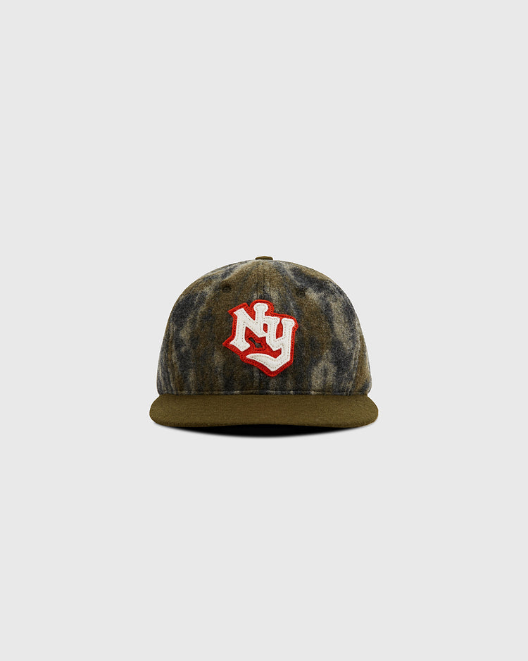 The Ebbets Field Mossy Oak x New York Knights Ballcap features a brown and gray camo pattern with a red and white NY logo on the front, displayed against a plain light gray background.